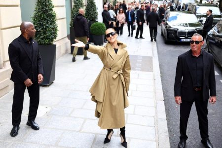 Celine Dion, in a tan trench coat and dark sunglasses, smiles and throws one arm out to greet fans on a Paris street outside her hotel, flanked by two security staff.