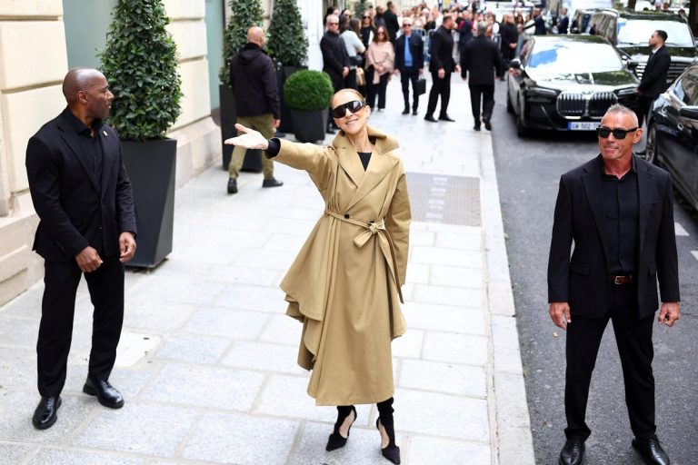 Celine Dion Returns to the Stage in Paris, Six Years After Stiff Person Syndrome Stopped Her Celine Dion, in a tan trench coat and dark sunglasses, smiles and throws one arm out to greet fans on a Paris street outside her hotel, flanked by two security staff.