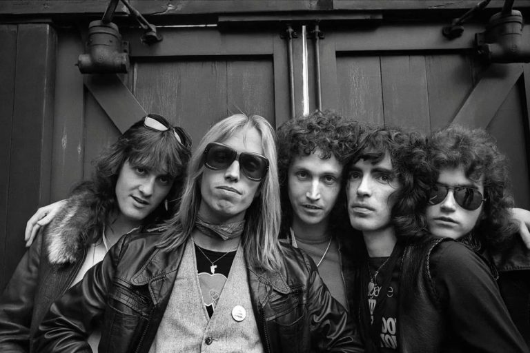 Black and white 1970s portrait of the five original members of Tom Petty and the Heartbreakers standing together against a wooden door, Tom Petty in sunglasses at the center