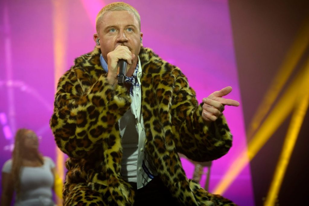 Macklemore Dropped From Ed Sheeran’s Loop Tour After ‘Free Palestine’ Speech at MetLife Stadium Macklemore performing onstage in a leopard print fur coat, holding a microphone to his mouth and gesturing with his right hand, against pink and gold stage lighting
