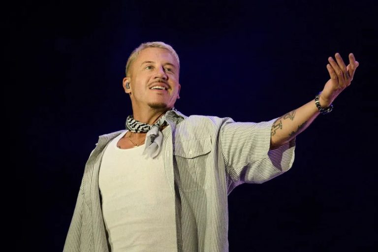 Macklemore Donates His Entire $1 Million Ed Sheeran Tour Fee – and Invites Robert Kraft to Match It Macklemore performing on stage with one arm outstretched, wearing a patterned scarf and a striped shirt against a dark backdrop