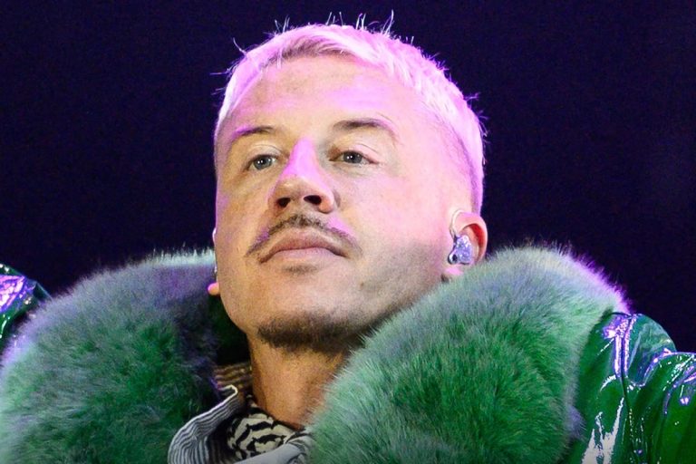 Close-up of Macklemore performing onstage under pink lighting, wearing a green fur-collared jacket