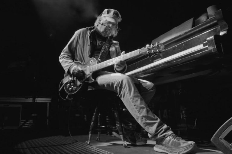 Neil Young seated on a stool playing a Les Paul electric guitar beside a grand piano on a darkened stage, photographed in black and white