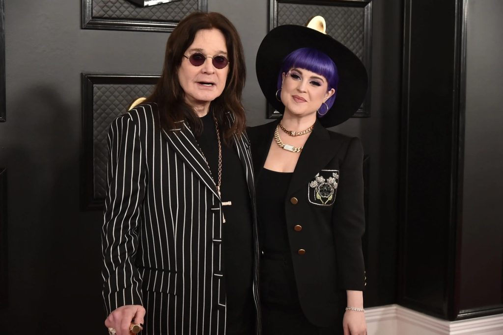 Ozzy Osbourne in a pinstriped suit and round sunglasses standing beside his daughter Kelly Osbourne, who wears a wide-brimmed black hat and purple hair, on a Grammy Awards red carpet