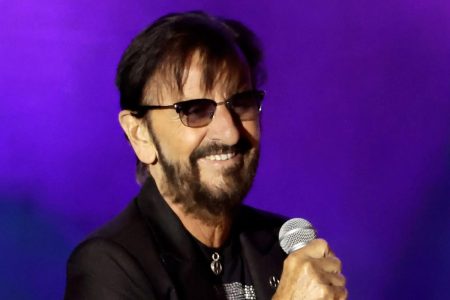 Ringo Starr Says the Beatles Would Have Got Back Together If Lennon and Harrison Had Lived Ringo Starr, wearing tinted glasses and a black jacket, smiles while holding a microphone against a purple backdrop