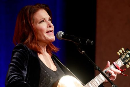 Rosanne Cash singing into a microphone on stage, playing an acoustic guitar, wearing a black velvet jacket against a blue-lit backdrop
