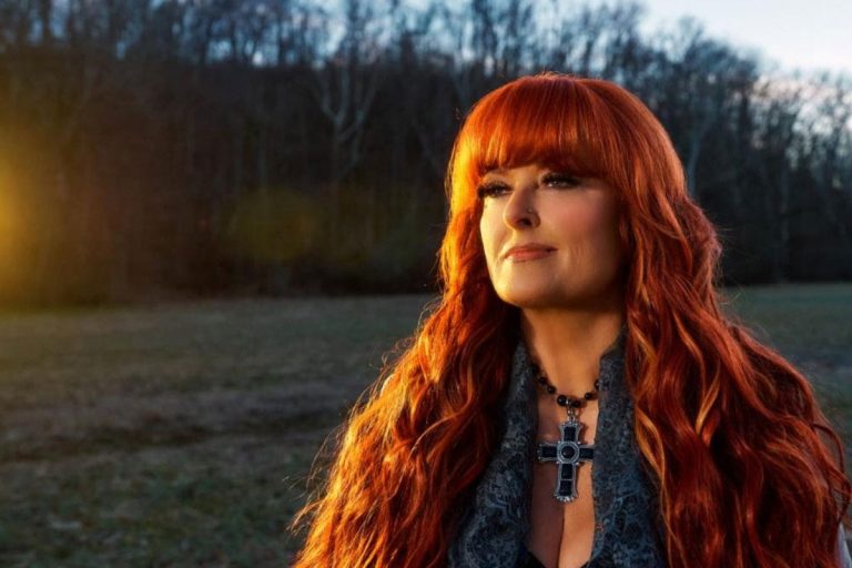 Wynonna Judd with long red hair and a cross pendant, photographed outdoors at sunset