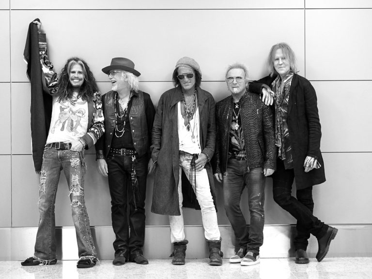 Black-and-white photo of the five members of Aerosmith leaning against a wall