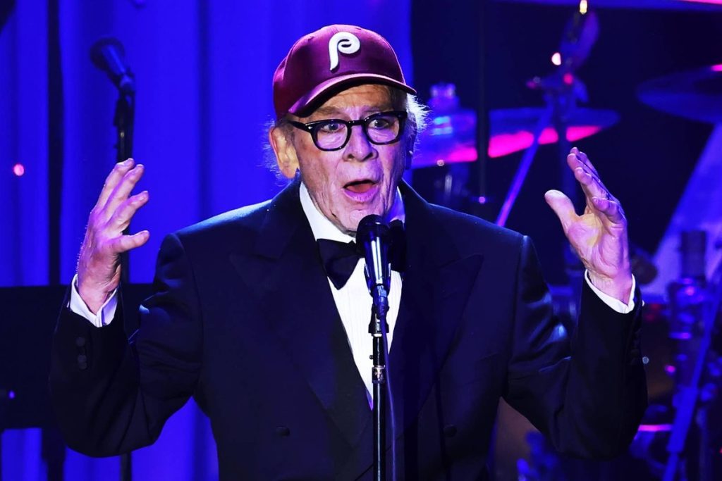 Art Garfunkel Postpones UK Tour After Respiratory Infection: ‘My Heart Is Broken’ Art Garfunkel in a tuxedo and red baseball cap singing at a microphone with his hands raised