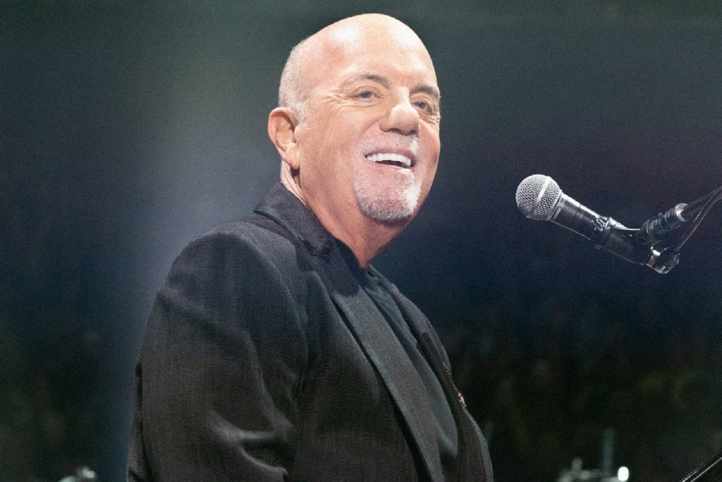 Billy Joel Says He Isn’t Allowed to Drive as Doctors Weigh Whether He Can Perform Again Billy Joel smiling at the piano microphone on stage in a dark jacket