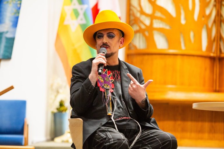 Boy George in a yellow hat speaking into a microphone at Temple Sholom in Vancouver