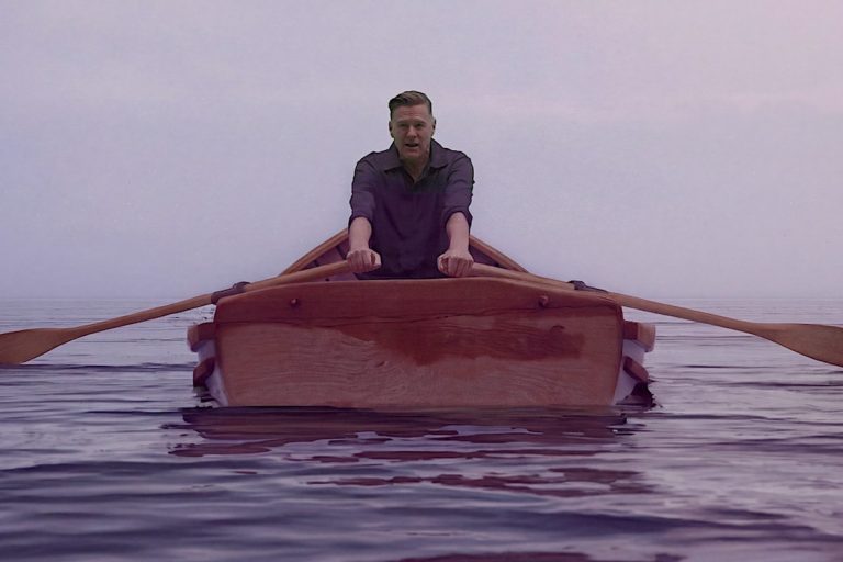 Bryan Adams rowing a wooden boat across calm water in the Lake Ontario music video