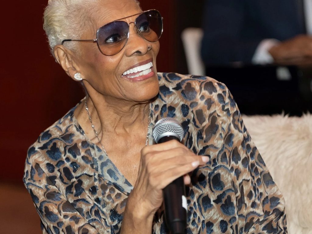 Dionne Warwick Cancels Carnegie Hall Appearance, Says Leg and Foot Pain Left Her Unable to Stand or Walk Dionne Warwick smiling in aviator sunglasses and a leopard-print blouse, holding a microphone
