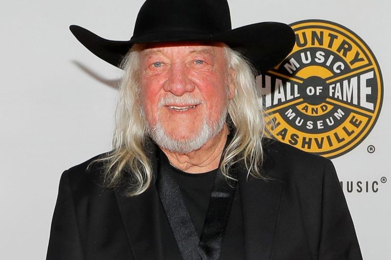 John Anderson in a black cowboy hat and black jacket smiling in front of a Country Music Hall of Fame and Museum backdrop