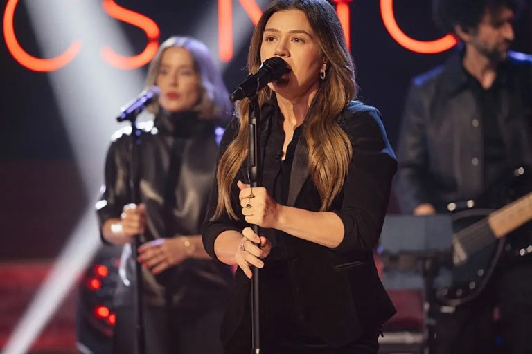 Kelly Clarkson singing into a microphone on stage in a black blazer, with a backing singer and guitarist behind her