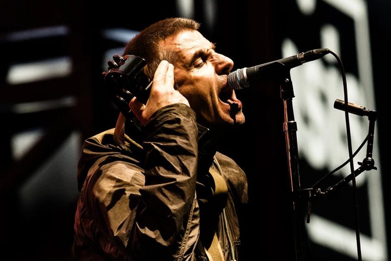 Liam Gallagher singing into a microphone on stage, holding a tambourine to his head