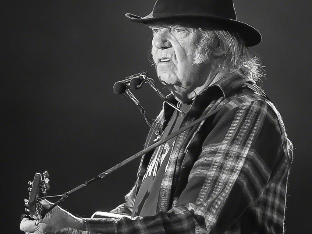 Neil Young in a black hat and plaid shirt, playing acoustic guitar with a harmonica rack on stage