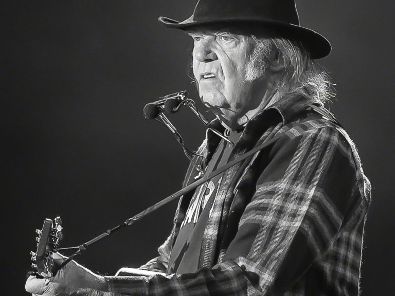 Neil Young Sues Trump PAC for $10 Million Over ‘Rockin’ in the Free World’ — Again Neil Young in a black hat and plaid shirt, playing acoustic guitar with a harmonica rack on stage