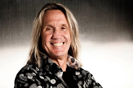 Iron Maiden’s Nicko McBrain Turned Down Golf With Trump, Says He and His ‘Cronies’ Should Be Jailed Nicko McBrain smiling in a black patterned shirt against a gray studio backdrop