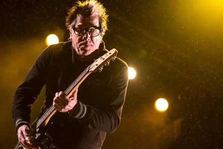 Noodles of The Offspring playing guitar on stage under yellow lights
