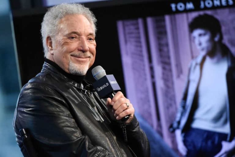 Tom Jones in a black leather jacket smiling while holding a microphone at a Q&A event