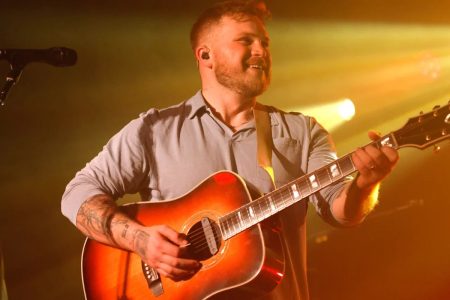 Zach Bryan smiling on stage playing a sunburst acoustic guitar under yellow stage lights